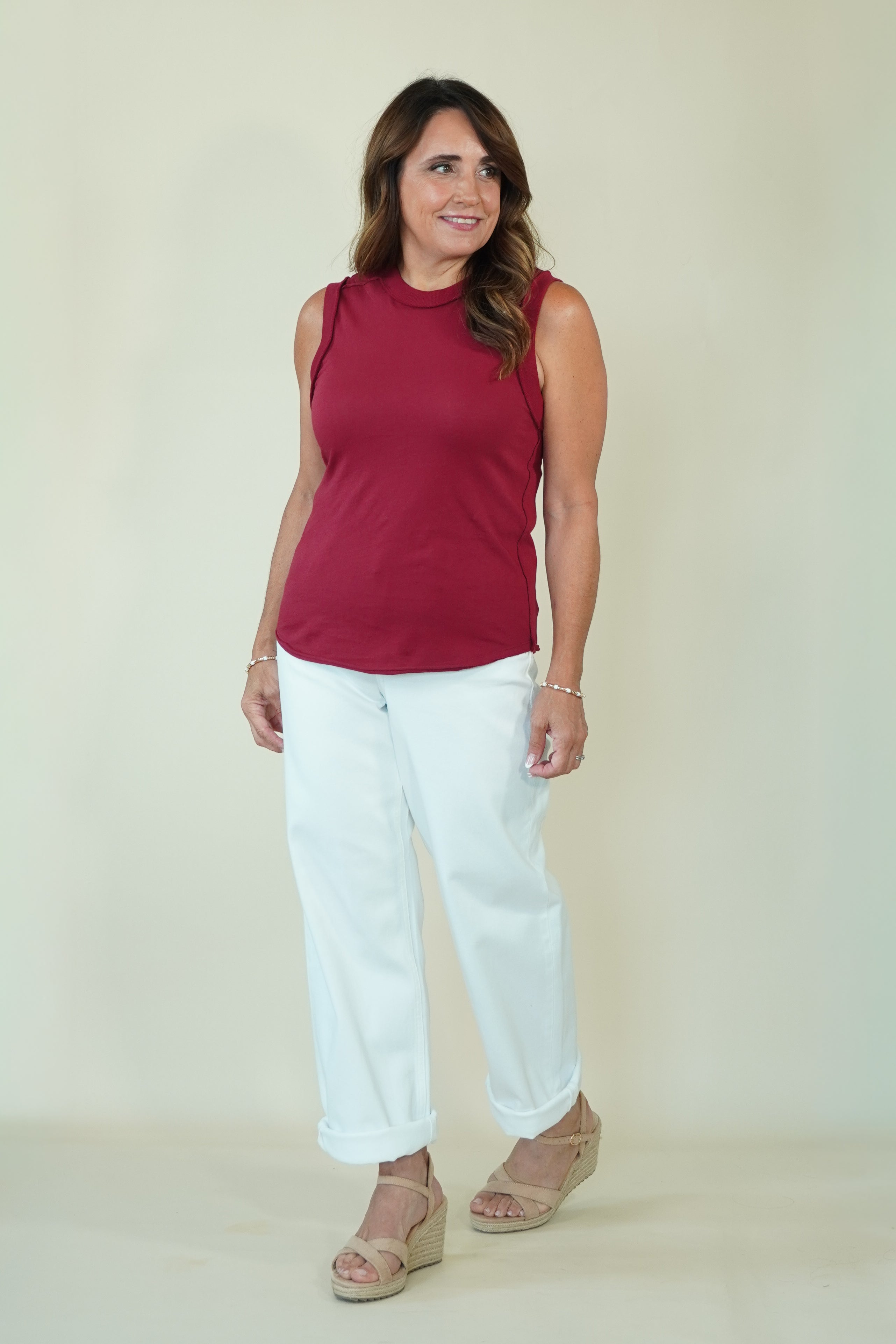 A woman modeling a sleeveless burgundy top and white cropped pants with beige wedge sandals, standing against a plain light beige studio background.