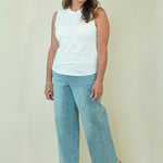 A woman modeling a white sleeveless top and light blue wide-leg cropped jeans with beige wedge sandals, standing against a plain cream-colored studio background.