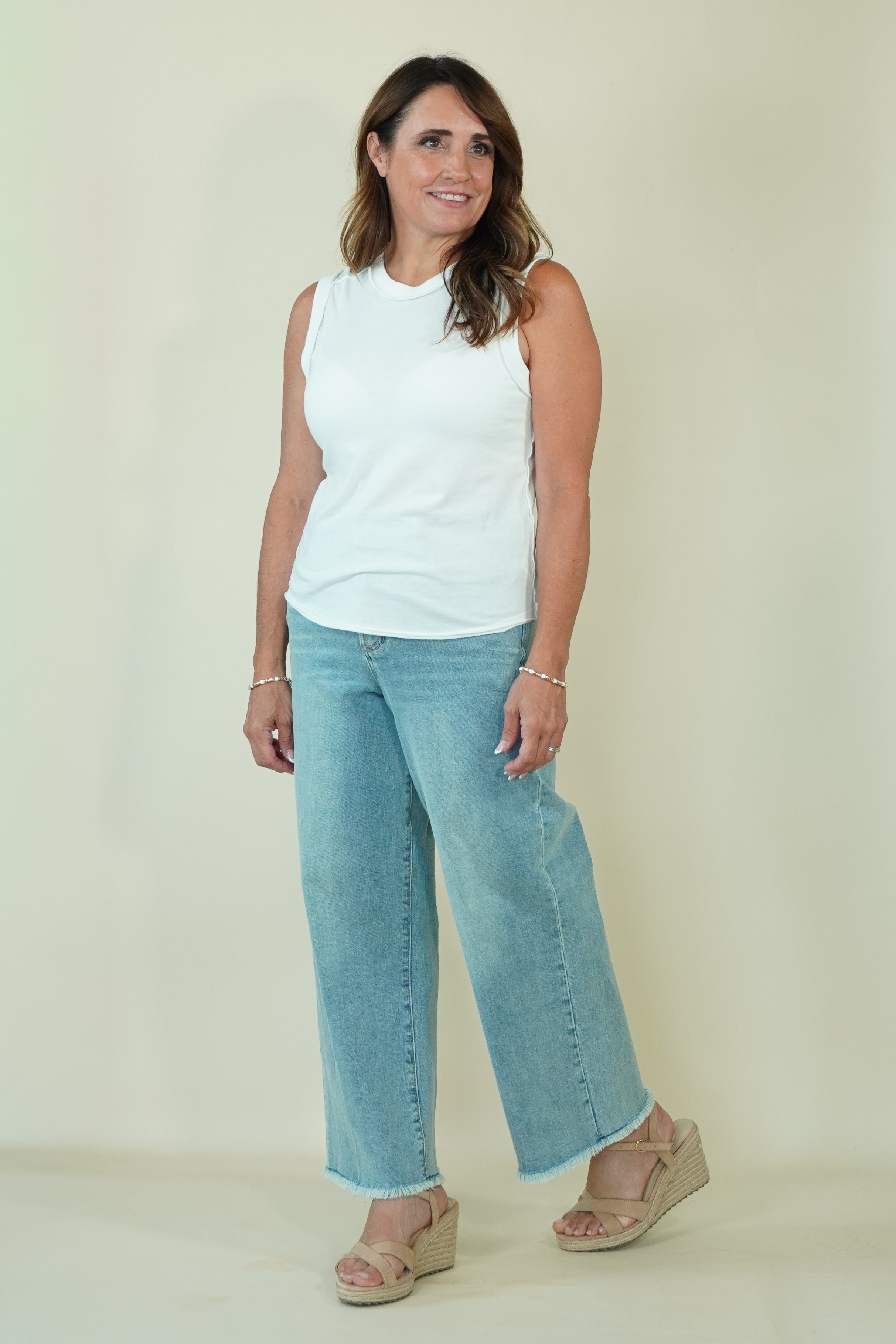 A woman modeling a white sleeveless top and light blue wide-leg cropped jeans with beige wedge sandals, standing against a plain cream-colored studio background.