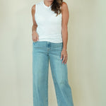 A woman modeling a white sleeveless top and light-wash wide-leg blue jeans against a plain beige studio background.