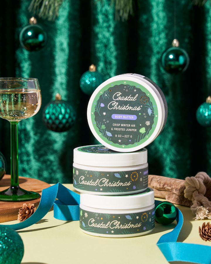 Three stacked tins labeled 'Coastal Christmas' with a glass of champagne and festive decorations in the background.