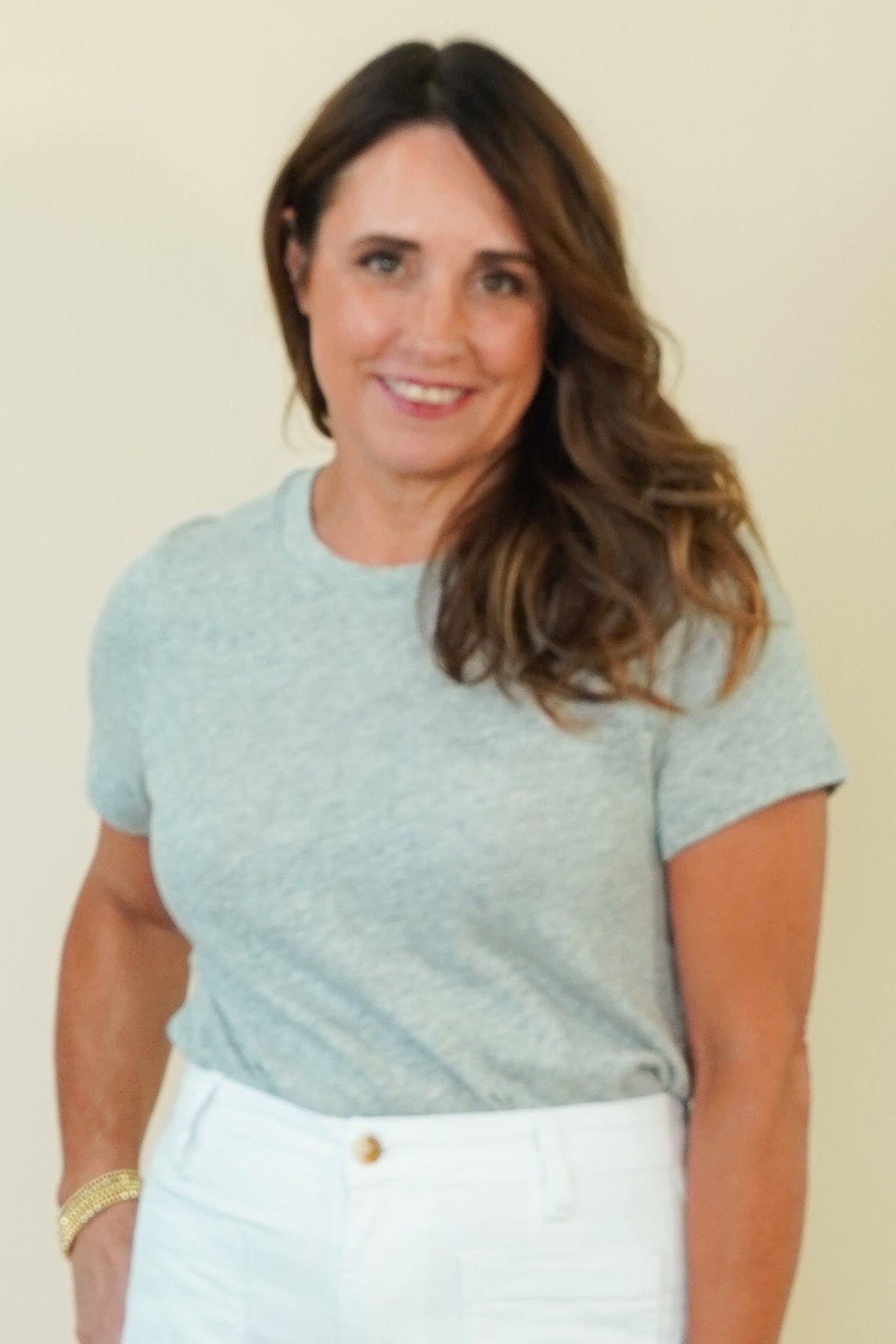 A woman modeling a light heather gray short-sleeve t-shirt and white denim shorts against a solid cream-colored studio background.