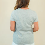 A woman viewed from behind wearing a light heather gray short-sleeve t-shirt and white shorts, standing against a plain beige background.