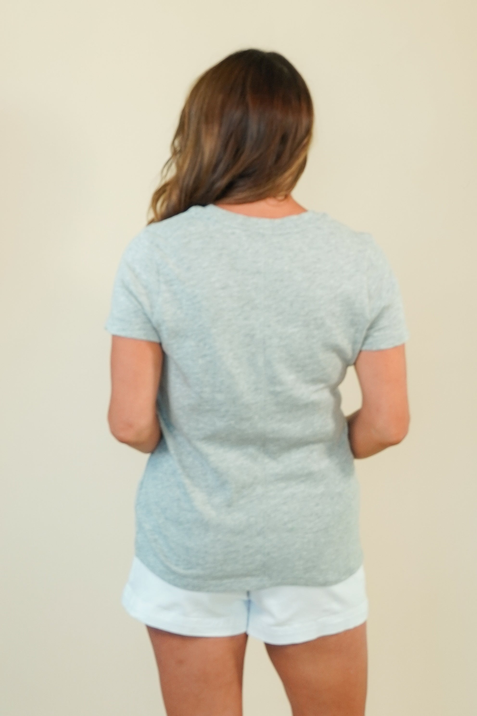 A woman viewed from behind wearing a light heather gray short-sleeve t-shirt and white shorts, standing against a plain beige background.