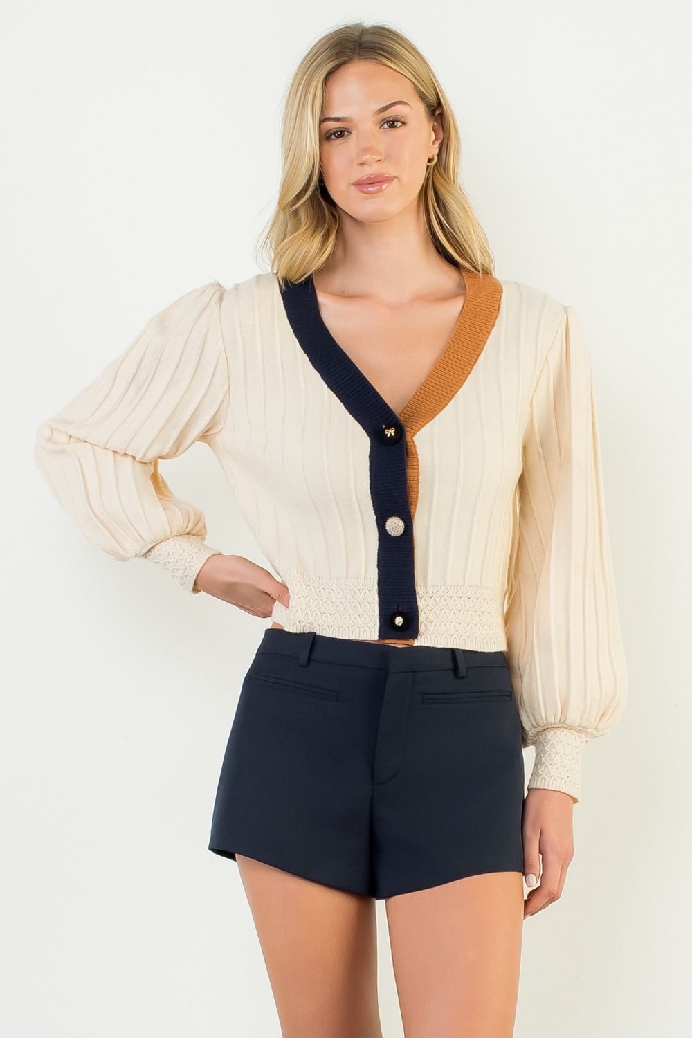 Woman wearing a cream cardigan with a deep neckline and navy buttons, paired with black shorts on a white background.