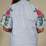 Person wearing a light blue shirt with floral embroidery and beige pants on a plain background