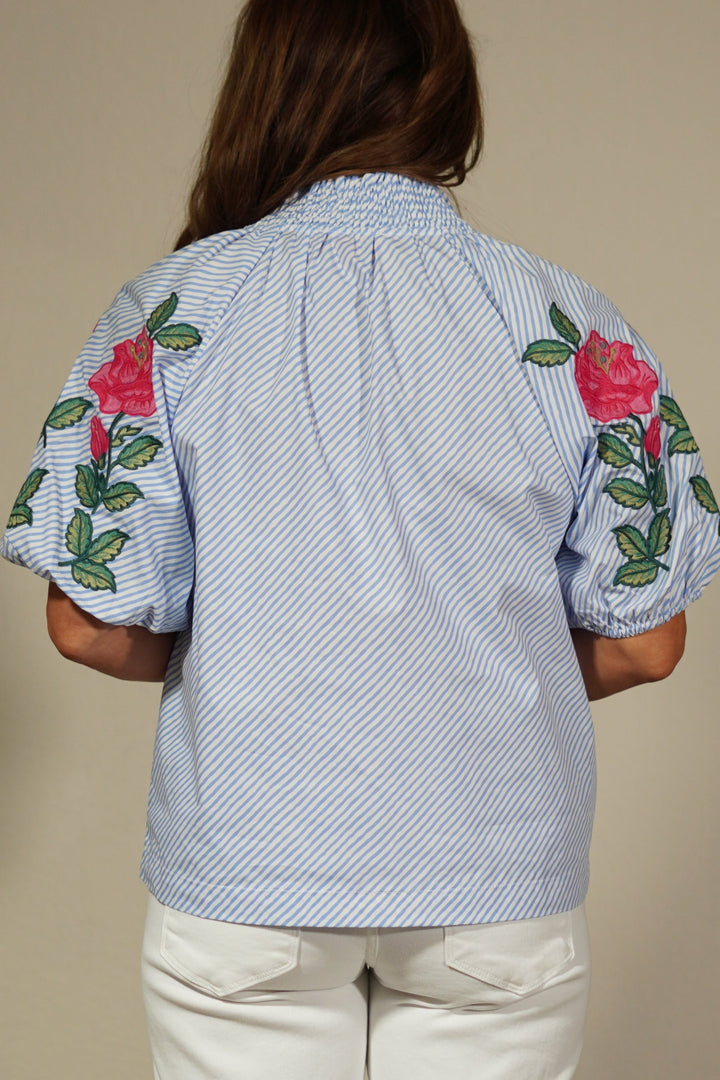 Person wearing a light blue shirt with floral embroidery and beige pants on a plain background