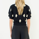 Person wearing a black sweater with white diamond patterns and black shorts on a white background