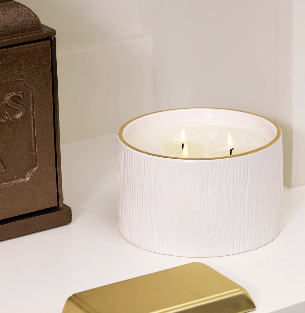 White candle in a textured container with a gold rim on a white surface, next to a brown box.