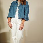 Woman wearing a blue denim jacket over a white dress, standing against a plain wall.
