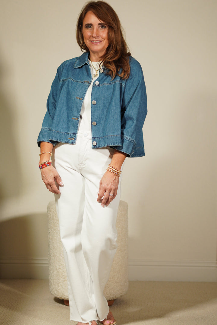 Woman wearing a blue denim jacket over a white dress, standing against a plain wall.