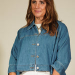 Woman wearing a blue denim jacket and white pants standing against a plain wall.