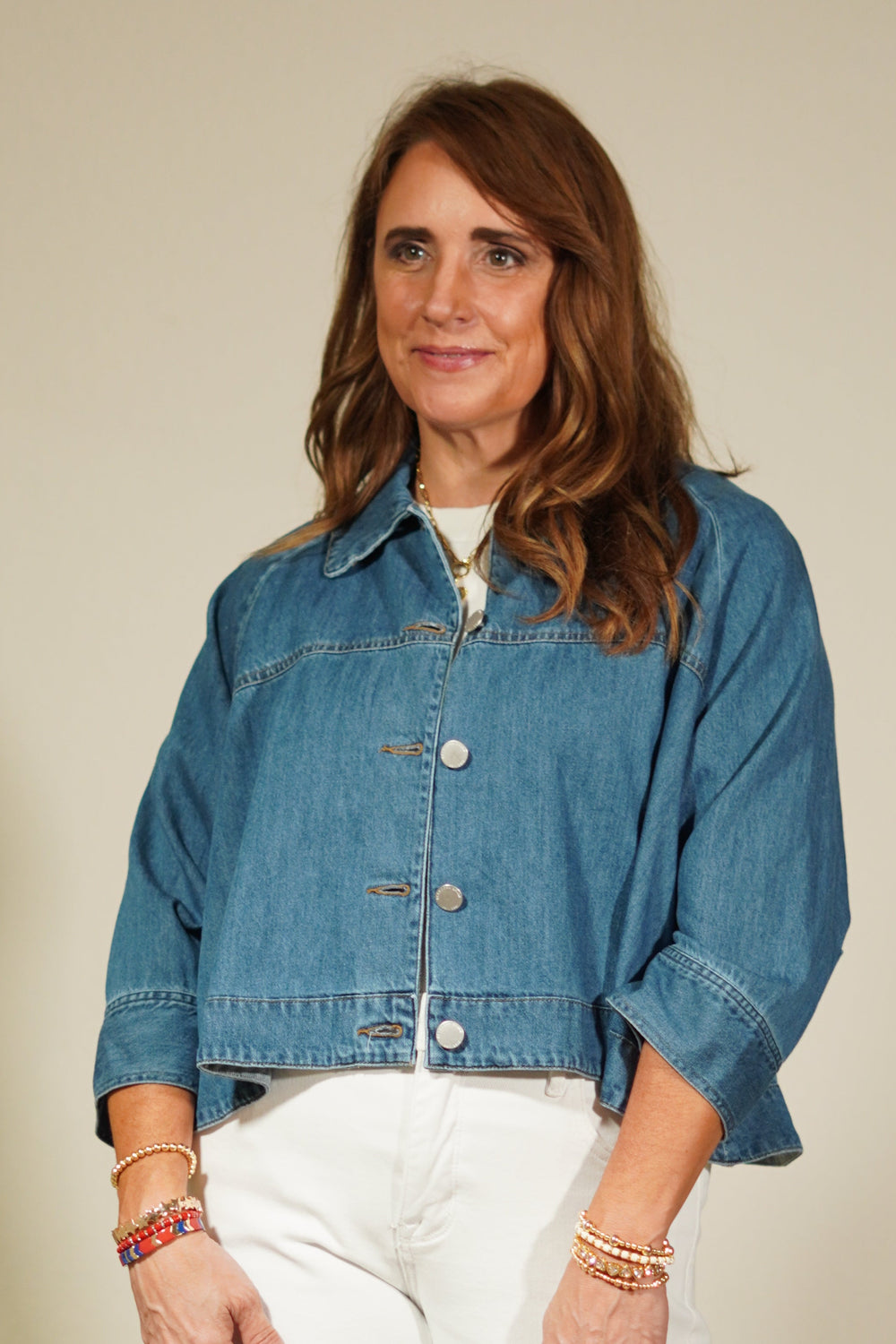 Woman wearing a blue denim jacket and white pants standing against a plain wall.