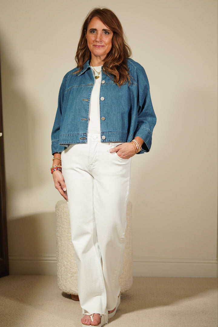Person wearing a blue denim jacket over a white outfit, standing against a plain wall.