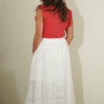Woman wearing a red top and white skirt against a plain wall.