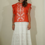 Woman wearing a red top with white floral patterns and a white skirt against a plain background