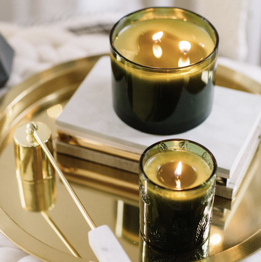 Two lit candles in black glass containers on a gold tray with a white surface.