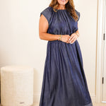 Woman in a blue dress standing in a room with a mirror and white dresser.