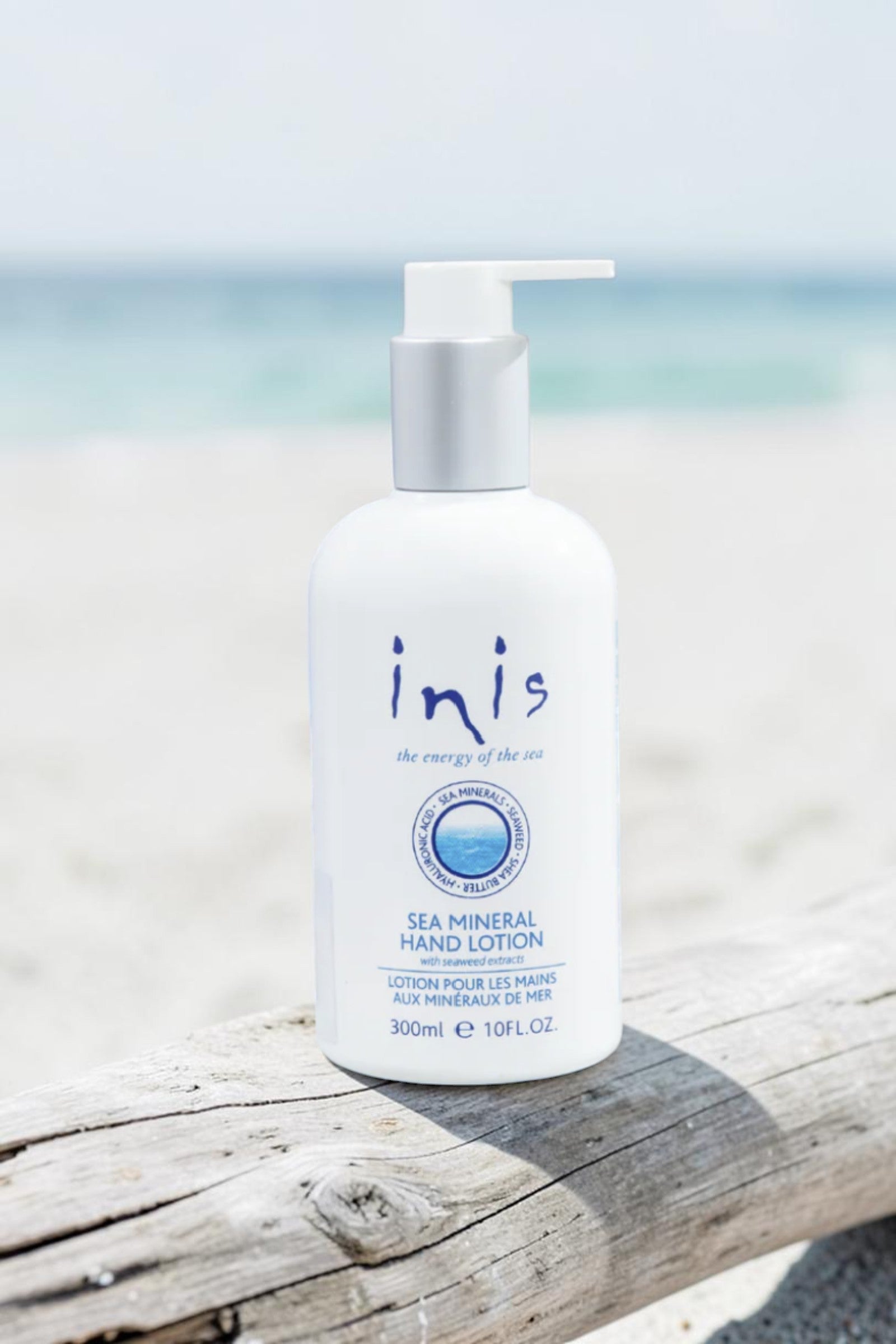 A 300ml bottle of Inis Sea Mineral Hand Lotion on a light marble surface, styled with a dollop of white lotion, a woven tray, and a blurred potted plant in the background.
