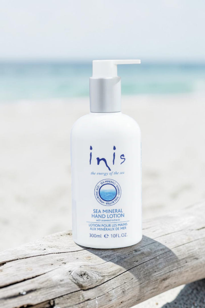 A 300ml bottle of Inis Sea Mineral Hand Lotion on a light marble surface, styled with a dollop of white lotion, a woven tray, and a blurred potted plant in the background.