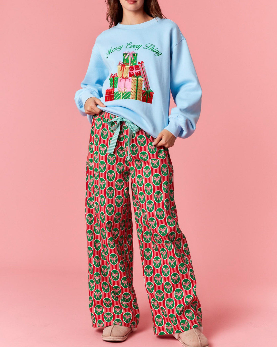 Woman wearing a light blue sweater with a Christmas design and red patterned pants on a pink background
