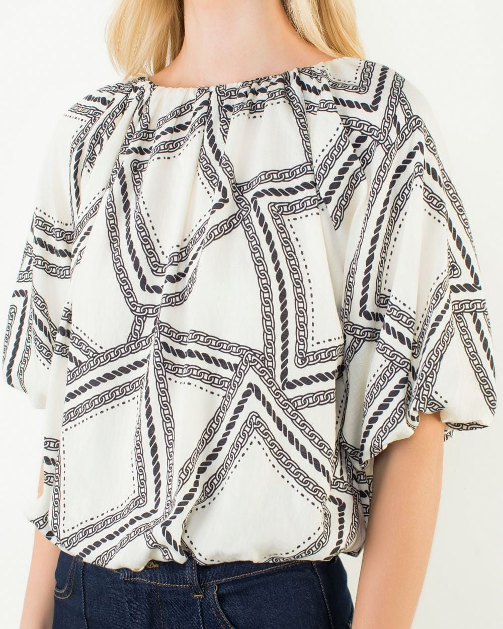 Woman wearing a white blouse with black geometric pattern and dark jeans on a plain background