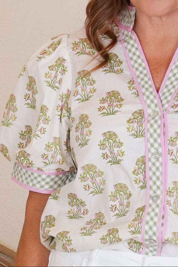 Woman standing in a room wearing a floral blouse and white pants.