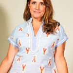 Woman wearing a light blue blouse with orange lobster pattern and orange pants against a beige background