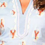 Woman wearing a light blue blouse with orange lobster pattern and orange pants against a beige background