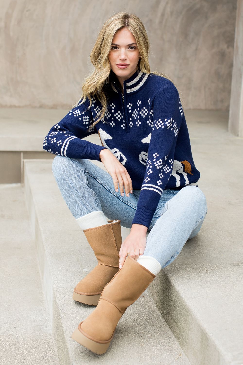 Woman wearing a blue sweater with white patterns, light blue jeans, and tan boots sitting on steps.
