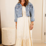 Woman standing in a room wearing a white dress and denim jacket.