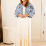 Woman standing in a bedroom wearing a denim jacket and white dress.