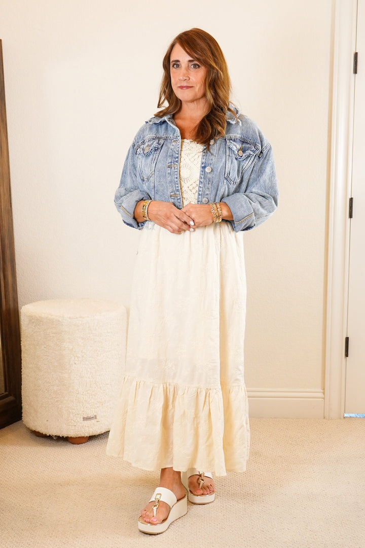 Woman standing in a bedroom wearing a denim jacket and white dress.
