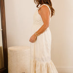 Woman in a white dress standing in a bedroom next to a dresser and mirror.