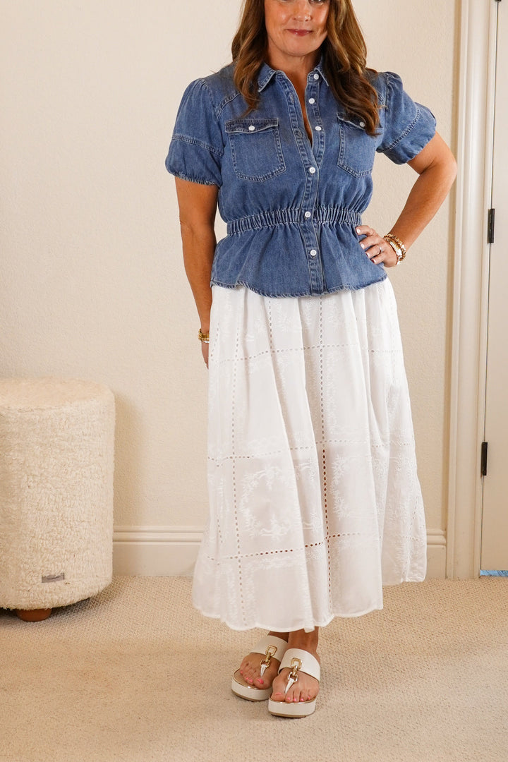 Woman wearing a blue denim shirt and white skirt in a bedroom.