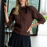 Woman wearing a brown cable knit sweater and black pleated skirt indoors.