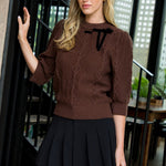 Woman wearing a brown cable knit sweater and black pleated skirt in an indoor setting.