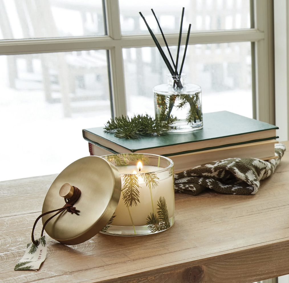 Candle with gold lid on a wooden surface with books and decorative items in the background.
