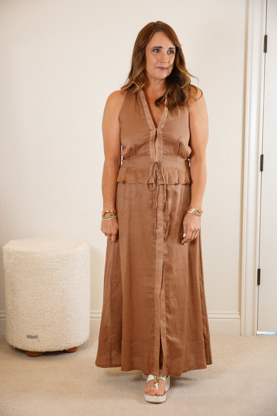 Woman wearing a brown sleeveless top and long skirt standing in a room with a white ottoman.