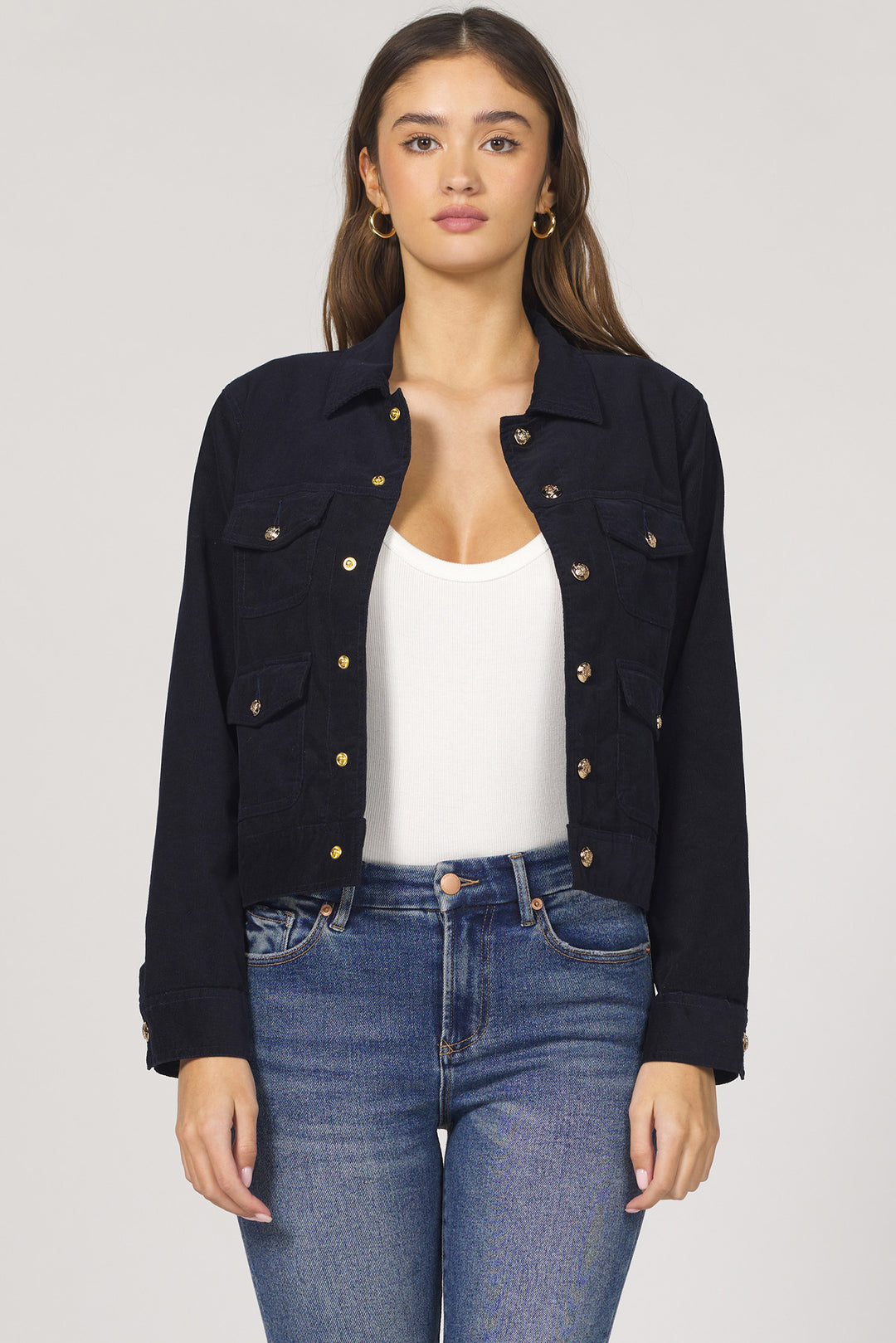Woman wearing a black denim jacket with gold buttons, white top, and blue jeans on a light gray background