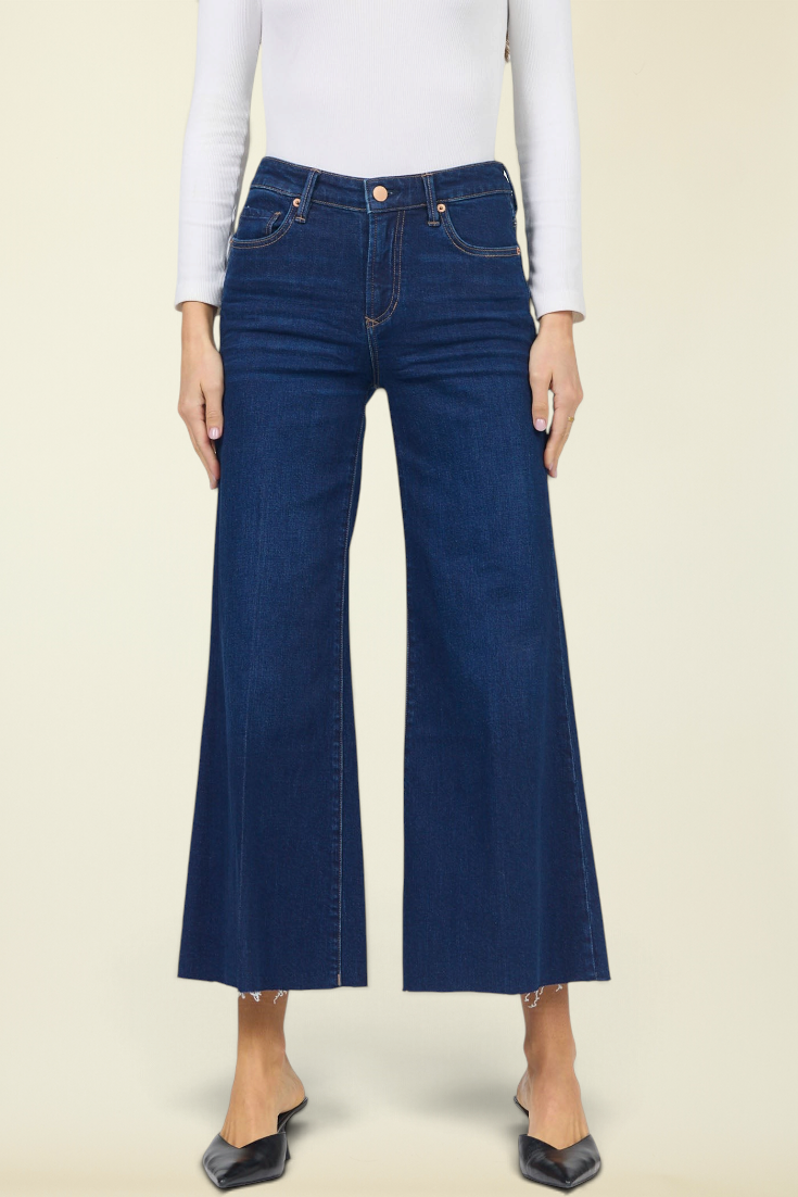 A model wearing dark blue wide-leg cropped jeans, a white long-sleeve top, and black pointed flats, standing against a solid light beige background.