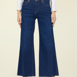 A model wearing dark blue wide-leg cropped jeans, a white long-sleeve top, and black pointed flats, standing against a solid light beige background.