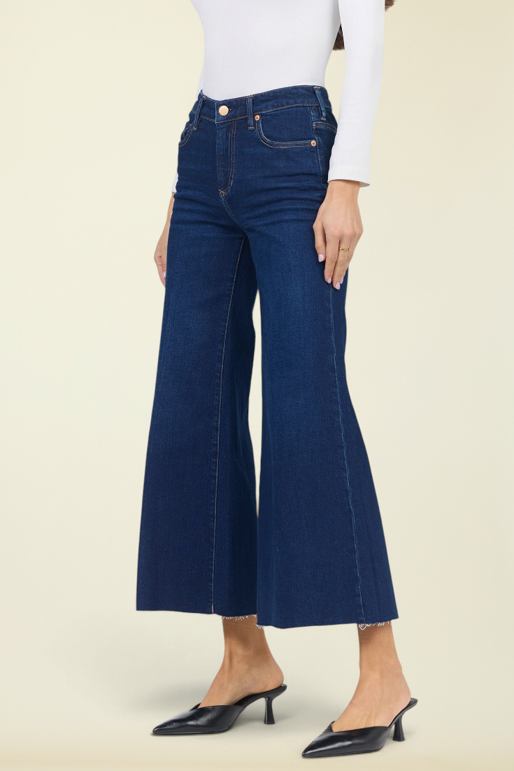 A model wearing dark blue wide-leg cropped jeans with a frayed hem, styled with a white long-sleeve top and black pointed-toe mule heels against a solid light beige background.