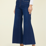 A model wearing dark blue wide-leg cropped jeans with a frayed hem, styled with a white long-sleeve top and black pointed-toe mule heels against a solid light beige background.