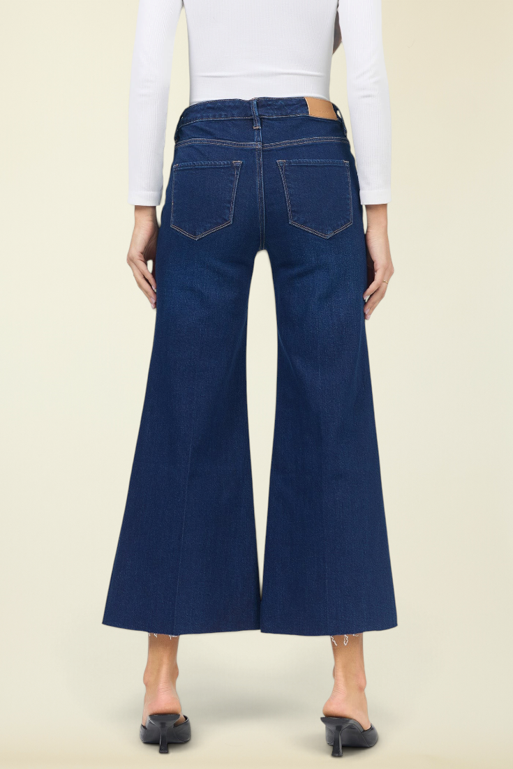 Back view of a model wearing dark blue wide-leg cropped jeans with a white long-sleeve top and black mule heels, set against a solid light beige background.