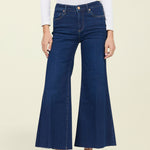 A woman modeling dark blue wide-leg cropped jeans with a white long-sleeve top and black pointed-toe mule heels, standing against a solid light beige background.
