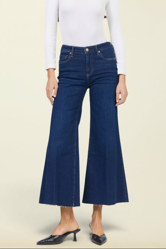 A woman modeling dark blue wide-leg cropped jeans with a white long-sleeve top and black pointed-toe mule heels, standing against a solid light beige background.