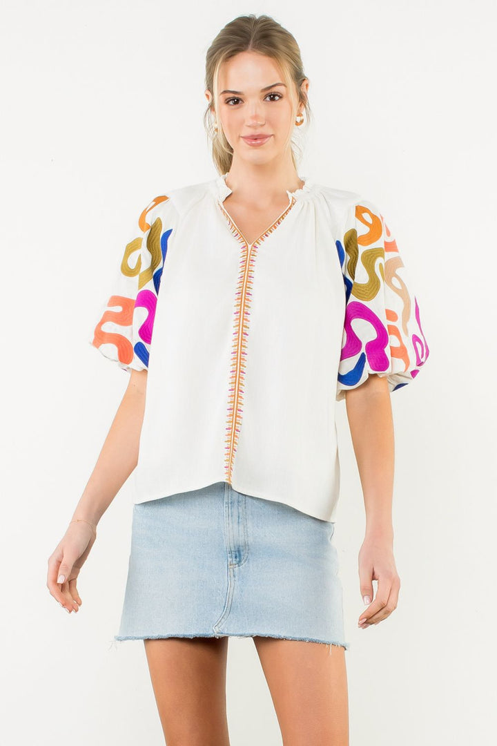 Woman wearing a white blouse with colorful patterns on sleeves and a denim skirt against a white background