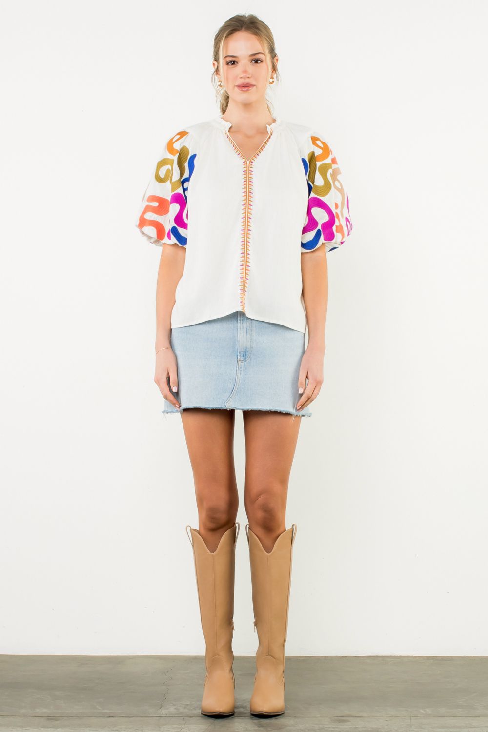 Woman wearing a colorful embroidered top, denim skirt, and tan boots on a white background