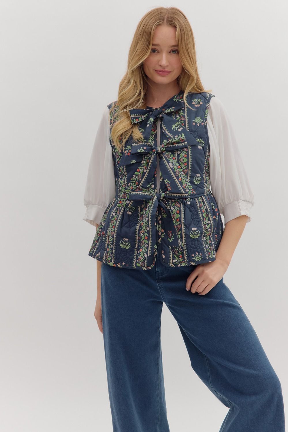 Woman wearing a patterned vest over a white shirt with blue pants on a white background
